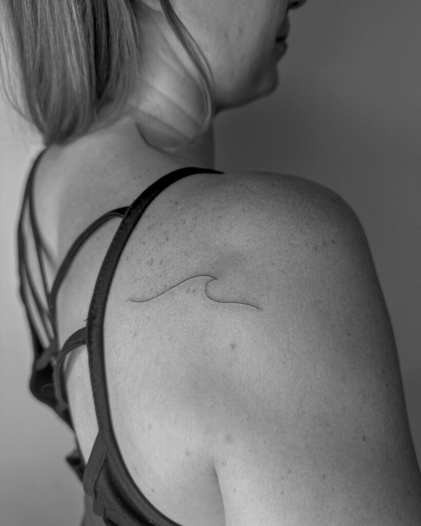 Delikatny, linearny tatuaż przedstawiający falę, wykonany na łopatce kobiety; minimalistyczny styl inspirowany naturą i fantastyką.